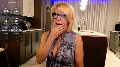 Media: Video of a blonde, middle-aged woman with glasses and a denim vest, covering her mouth in a modern kitchen.