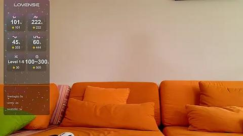 Media: A video of a minimalist, modern living room featuring a large orange sofa with orange cushions against a plain white wall. A weather app display is visible in the left corner.