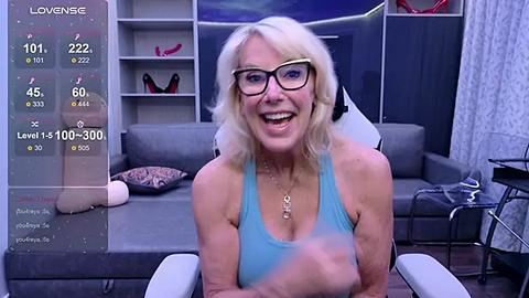 Media: Video of a middle-aged blonde woman with glasses, wearing a blue tank top, sitting on a gray couch in a modern living room.