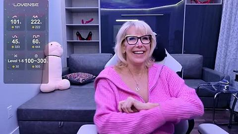 Media: Video of a smiling, middle-aged blonde woman with glasses, wearing a pink sweater, sitting on a grey couch in a modern living room.