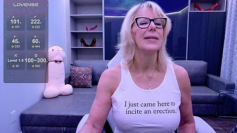 Media: Video of a blonde woman with glasses wearing a white tank top with \"I just came here to insert an erection\" printed on it, in a modern living room.