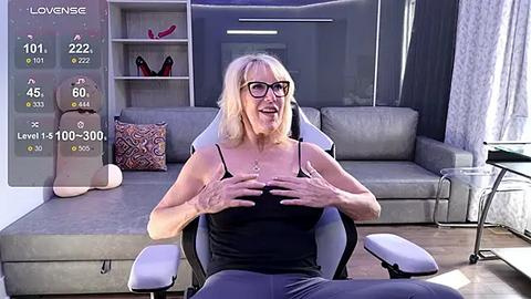 Media: Video of a middle-aged woman with blonde hair and glasses, sitting in a modern gray leather chair, wearing a black tank top, holding her breasts. Background includes a gray sofa with patterned cushions, a wooden floor, and a digital screen displaying stats.