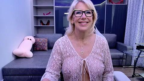 Media: Video of a smiling, blonde woman with glasses, wearing a light pink lace cardigan, seated on a gray sofa in a modern living room with shelves, a plush toy, and a decorative pillow.