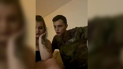 Media: A video shows a young woman with light skin and long blonde hair, wearing a black top, leaning on a man in camouflage attire. They sit on a beige sofa in a dimly lit room.