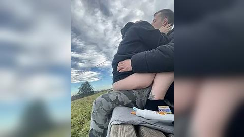 Media: Video of a couple kissing passionately outdoors on a bench, surrounded by a cloudy sky and green grassy landscape. The man, in camo pants and a black hoodie, lifts the woman in a black dress, revealing her bare legs.