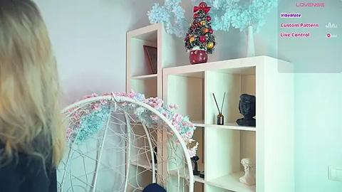 Media: Video of a light-skinned woman with blonde hair, standing in a white-walled room with a wooden shelving unit. The shelves hold a decorative Christmas tree, a Buddha statue, and other decorative items. A white, wire-framed hanging chair is in the foreground.