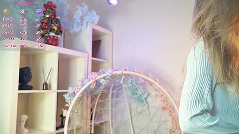 Media: Video of a woman in a light blue sweater, standing in a room with a white shelving unit, a small tree, and a large, decorative, floral birdcage.