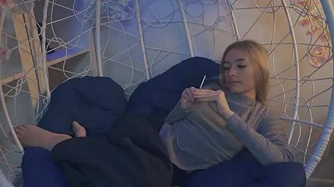 Media: Video of a young blonde girl in a gray sweater and black pants, seated in a white wicker swing with a dark blue cushion, reading a book, indoors.