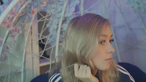 Media: Video of a young woman with long, blonde hair, looking contemplative, adjusting her hair near a pink and white floral parasol in a softly lit room.