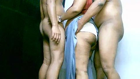 Media: Video of three nude individuals, two males and one female, standing closely. The female, with light skin, wears white panties. The males, with muscular builds, are partially visible. Background is dark and indistinct.