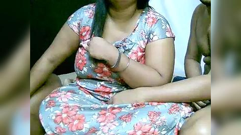 Media: Video of a South Asian woman with medium skin tone, long black hair, wearing a floral dress, sitting cross-legged, with a man's hand on her shoulder.