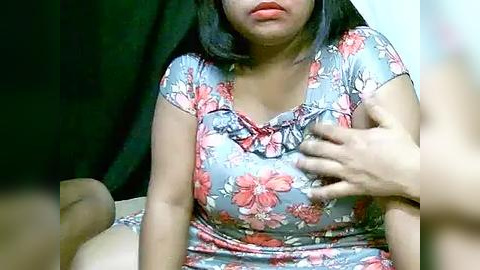 Media: Video of a young Asian woman with medium skin tone, shoulder-length black hair, wearing a floral-patterned top, holding her chest, surrounded by dark curtains, possibly indoors.