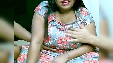 Media: Video of a South Asian woman with medium skin tone, long black hair, wearing a floral-patterned dress, sitting on a bed, smiling, with a hand on her chest, blurred background.