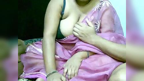 Media: Video of a partially clothed woman in a pink sari, exposing her left breast, with a man's hand covering her nipple. Background is dark and blurry.