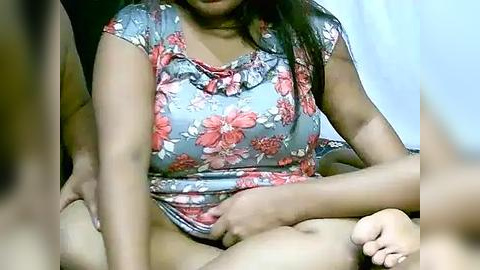 Media: A video captures a woman with medium brown skin, long dark hair, and a floral dress, sitting cross-legged with another person's hand on her thigh, creating a suggestive intimate scene.