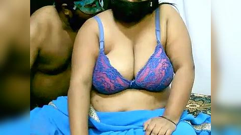 Media: Video of a plus-sized woman with medium brown skin, wearing a blue lace bra with pink accents and a blue skirt, sitting on a bed.