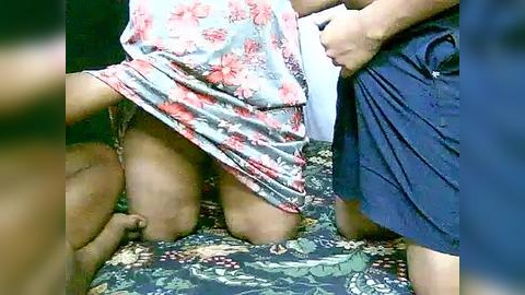 Media: Video of a man and woman kneeling on a floral-patterned bedspread, the woman in a floral dress, the man in dark shorts, arms wrapped around her.