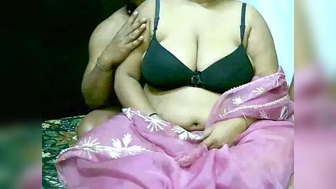 Media: Video of a middle-aged, overweight woman with medium skin tone, wearing a dark green bra and pink floral-patterned sari, sitting cross-legged on a dark green fabric.