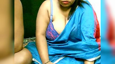 Media: Video of an East Asian woman with medium build, fair skin, and long black hair, wearing a blue sari with a pink lace bra visible, sitting on a dark background, blurry foreground.