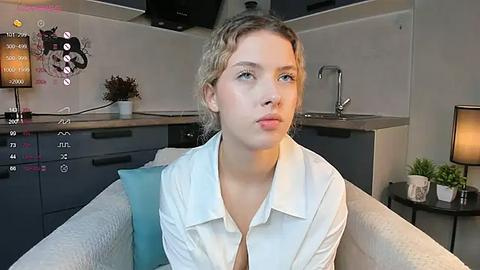 Media: Video of a young woman with blonde hair, wearing a white button-up shirt, seated in a modern, light-gray couch. Background includes a kitchen with dark cabinets, a sink, and a potted plant.