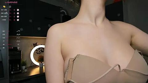 Media: Video of a woman in a beige bra, standing in a dimly-lit kitchen with a ring light, dark cabinets, and a potted plant.