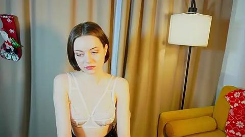 Media: A video of a young woman with straight, shoulder-length brown hair wearing a sheer white bra, sitting indoors against beige curtains. She has fair skin, is slender, and appears contemplative. Background includes a mustard-yellow armchair and a floor lamp with a white shade.