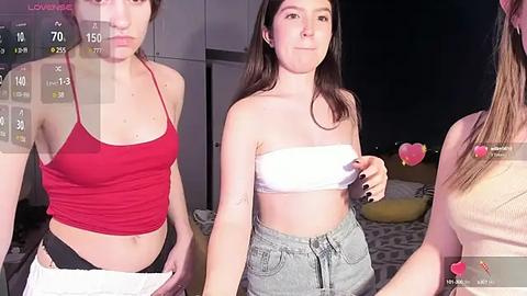 Media: Video of three women in a modern living room. Two are topless, one in a red tank top, another in a white strapless bandeau. One has gray sweatpants. Background features a dark room with minimal lighting.