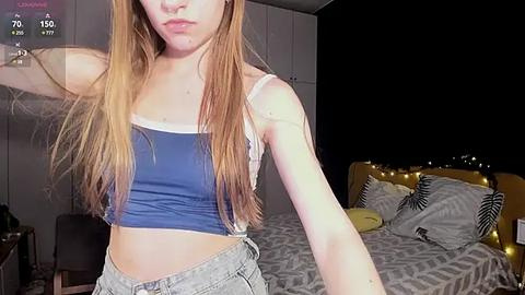 Media: Video of a young, fair-skinned woman with long, straight, blonde hair, wearing a blue crop top and light gray shorts, standing in a dimly lit bedroom with a bed and nightstand visible.