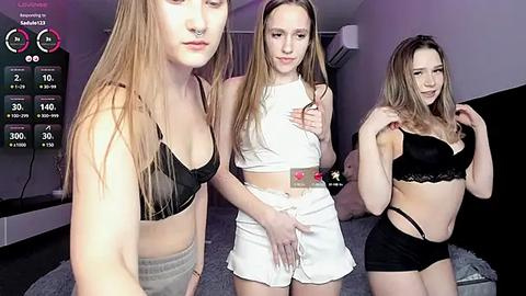 Media: Video of three young women in lingerie: one blonde in a black bra, one brunette in a white crop top, and one blonde in black lingerie. Bedroom background.