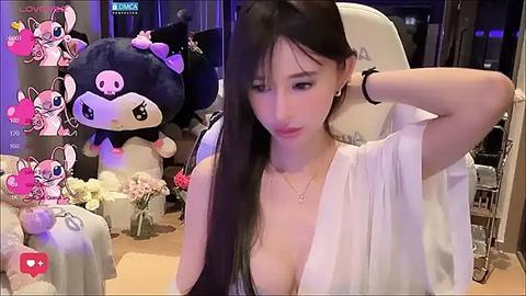 Media: A video of a young East Asian woman with long black hair, wearing a low-cut white blouse, seated in a cozy room adorned with plush toys and flowers.