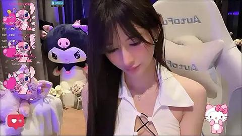 Media: Video of a young Asian woman with long black hair, wearing a white halter top, sitting in a plush chair. Background features plush toys and a \"Hello Kitty\" poster.