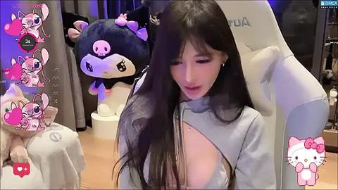 Media: Video of a young Asian woman with long black hair, wearing a revealing gray top, sitting in a plush chair with a large plush toy behind her.