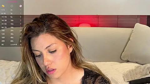 Media: A video of a young woman with light skin and long, wavy brown hair, wearing a black lace top, sitting on a beige couch in a modern, minimalist bedroom with a wooden headboard and red accent lighting.