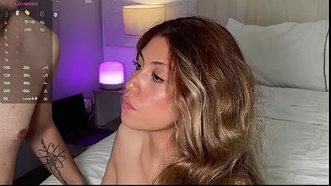 Media: Video of a young woman with long, wavy brown hair, light skin, and a small tattoo on her forearm, gazing at a screen displaying a virtual reality game. She's in a modern bedroom with a white bed, a nightstand, and a lamp.