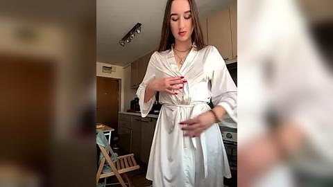 Media: A video of a young woman in a white satin robe, standing in a modern kitchen with wooden cabinets and stainless steel appliances, holding a small object.