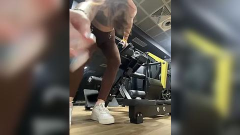 Media: Video of a woman in a gym, wearing dark leggings and white sneakers, adjusting a weight machine. Background shows gym equipment and a yellow towel on a bench.