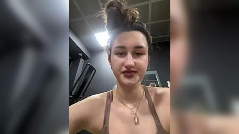 Media: A video of a young woman with fair skin, dark hair styled in a messy bun, wearing a brown tank top and gold necklace, in a gym setting with dim lighting.