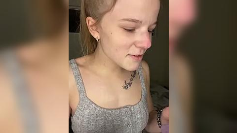 Media: A video of a young woman with fair skin, light brown hair pulled back, wearing a gray tank top with a small tattoo on her collarbone, standing in a dimly lit room.