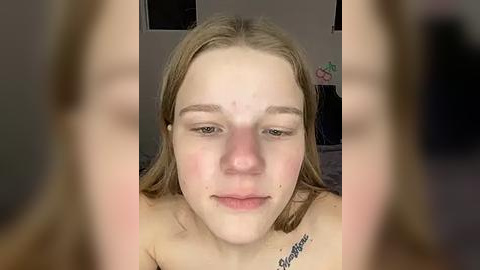 Media: Video of a young, fair-skinned, blonde woman with light makeup, slight acne, and a tattoo on her shoulder, smiling. Background shows a dimly lit room with a bed and indistinct objects.