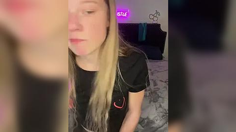 Media: Video of a young woman with long blonde hair, wearing a black shirt with a smiley face, sitting on a gray bed in a dimly lit bedroom with purple lighting and minimalist decor.