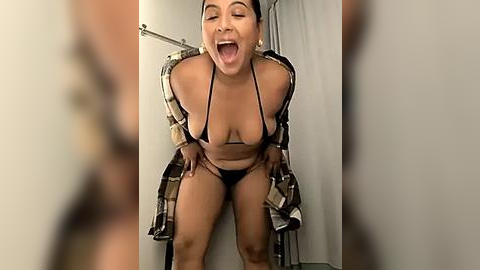 Media: A video of a curvy, dark-skinned woman in a revealing black bikini, with an open plaid shirt, leaning forward in a changing room, smiling widely.