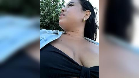 Media: Video of a young Asian woman with medium brown skin, wearing a black off-shoulder top, and a denim jacket, gazing up at a tree in the background.