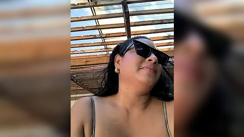 Media: Video of a smiling, light-skinned woman with long black hair, wearing sunglasses, a green tank top, and small hoop earrings, standing under a rustic, wooden-roofed structure.