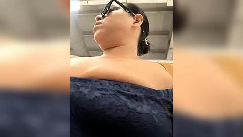 Media: Video of a woman with glasses and dark hair, wearing a navy blue lace bra, captured from a low angle, emphasizing her large breasts.