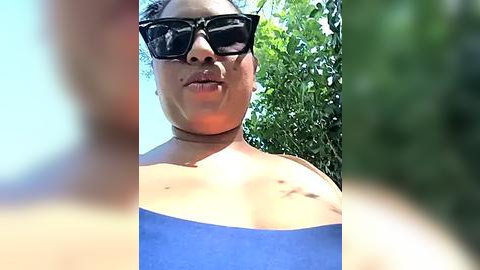 Media: Video of a person with medium-dark skin, wearing black sunglasses, a blue strapless top, and a choker necklace. They stand in a sunlit garden with green foliage. The image is slightly out of focus, with a blurred background.