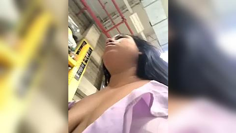 Media: Video of a young woman with medium-dark skin, long black hair, and a large breast exposed, wearing a light purple blouse. She is in an industrial setting with yellow warning signs and red pipes in the background.