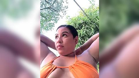 Media: A video of a Latina woman with medium skin tone, dark hair tied back, wearing an orange bikini top, captured from a low angle, with blurred green foliage in the background.