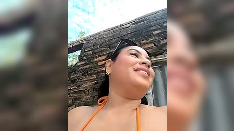 Media: Video of a smiling woman with long dark hair in a bright orange bikini, standing in front of an old, weathered brick wall.