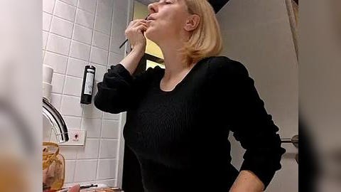 Media: Video of a woman with blonde hair, wearing a black sweater, brushing her teeth in a bathroom with white tiles and a sink.