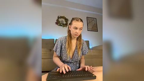 Media: Video of a young, fair-skinned woman with long, blonde hair in a floral dress, typing on a computer in a modern living room with beige furniture and minimalist decor.